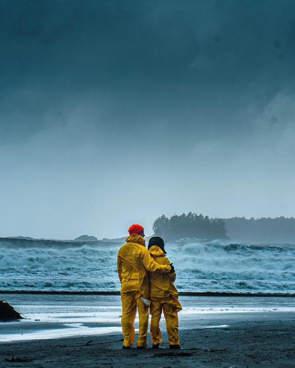 A couple in bright yellow raincoats stand on a blue monochrome landscape at the waterfront Tofino.
